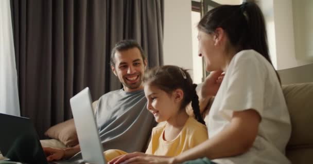 Family sitting on the sofa and looking at something on a gray laptop. A brunette girl in a white T-shirt, her daughter, a girl in a yellow dress, and a brunette man in a gray T-shirt are sitting - Filmati, video
