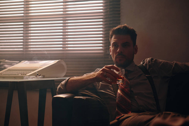 Mladý seriózní podnikatel v retro formalwear drží sklenici whisky, zatímco sedí na pohodlném gauči proti oknu s žaluziemi - Fotografie, Obrázek