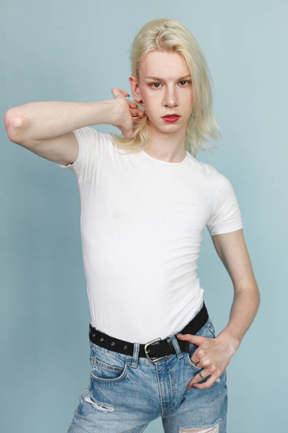 Young blonde man with make up on his face posing for a photo. Man wearing fashionable white t-shirt and has red lips. Man isolated on blue background. - Photo, Image