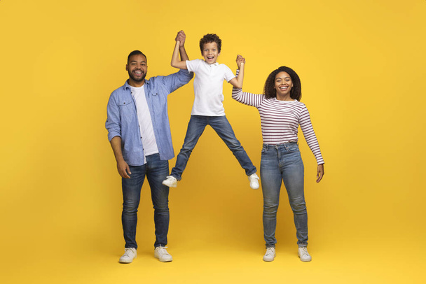 Cheerful Young Black Family Of Three Having Fun Together Over Yellow Studio Background, Happy African American Parents Holding Hands And Lifting Their Joyful Preteen Son, Bonding And Laughing - Фото, изображение