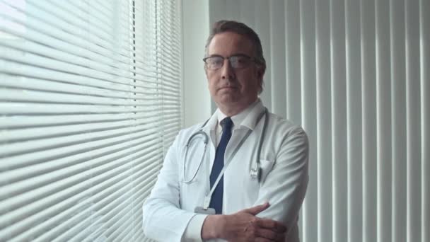 Portret starszego terapeuty w białym płaszczu w okularach patrzącego w kamerę, stojącego przy oknie z żaluzjami w klinice - Materiał filmowy, wideo