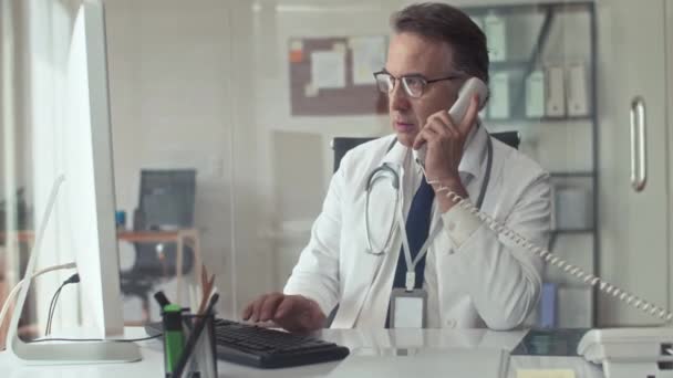 Tiro médio de terapeuta do sexo masculino preenchendo cartão médico no computador enquanto conversa com o paciente no telefone fixo na clínica - Filmagem, Vídeo