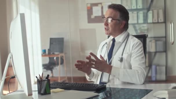 Μεσαία λήψη του ηλικιωμένου θεραπευτή καλώντας τον ασθενή σε βίντεο και συνταγογράφηση χάπια κάθεται μπροστά από τον υπολογιστή στην κλινική - Πλάνα, βίντεο