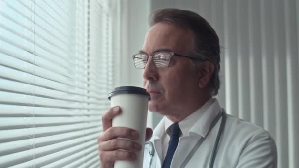 Middelmatige close-up van oudere arts in witte jas en glazen die koffie drinken terwijl ze rusten na een operatie in het ziekenhuis - Video