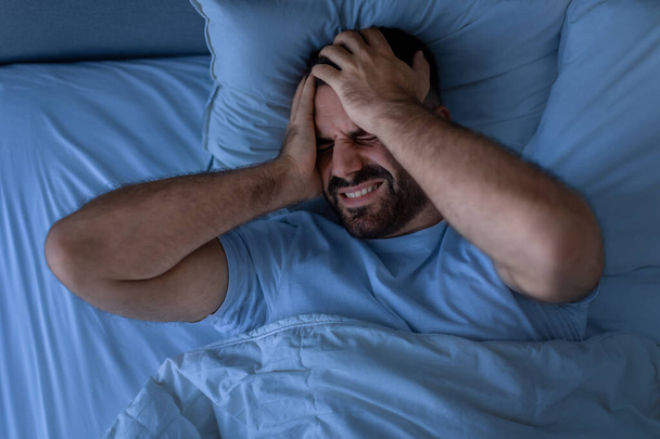 Schlaflosigkeit und Kopfschmerzen. Unglücklicher Typ, der an Migräne leidet und sich nachts krank fühlt, im Bett im modernen Schlafzimmer liegend, Aufnahme von oben, wie der Mann im schlechten Schlaf den Kopf berührt. Wenig Licht - Foto, Bild