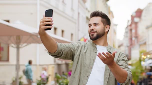 Bearded uomo blogger scattare selfie su smartphone, comunicare videochiamata online con gli abbonati o amici di famiglia, la registrazione di storie per i social media vlog, agitando ciao. Ragazzo in città urbana strada - Filmati, video