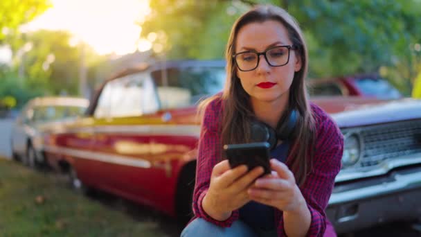 Donna con le cuffie è accovacciato accanto alla macchina d'epoca rossa e utilizzando smartphone sulla strada verde nel giorno d'estate. Tendenza alla moda.  - Filmati, video