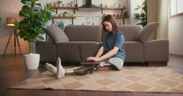 Giovane donna bruna con disabilità utilizza il computer portatile per digitare il piano di avvio del progetto. La donna completamente impegnata nel compito si siede sul pavimento a lavorare su gadget moderno - Filmati, video