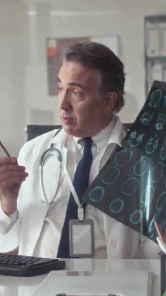 Pionowe ujęcie dojrzałego radiologa w białym płaszczu omawiającego rezonans magnetyczny z kolegą podczas rozmowy wideo w klinice - Materiał filmowy, wideo