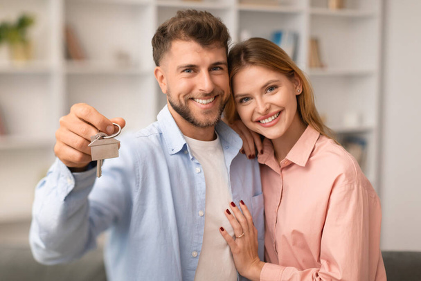Happy young caucasian couple holding home keys and hugging smiling at camera, romantic millennial family buying real estate property, man and woman poising in living room after moving - Photo, Image