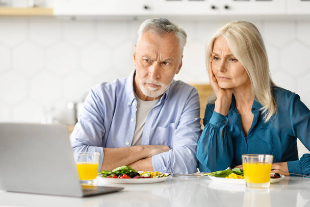 Беспокойная пожилая пара читает новости на ноутбуке во время завтрака на кухне, подчеркивает пожилые супруги, глядя на экран компьютера с озабоченностью, проверка входящей электронной почты, страдают проблемы, крупным планом - Фото, изображение