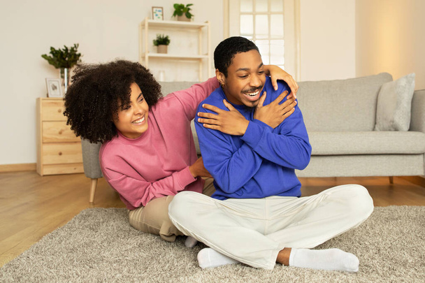 Giovane coppia afroamericana giocosamente solletico e ridere insieme nel loro interno casa, seduto sul pavimento, mostrando gioia e profondo legame romantico. Romanticismo giocoso nel matrimonio - Foto, immagini
