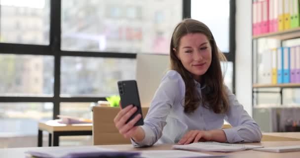 Genç kadın sekreter şirket ofisinde cep telefonuyla poz değiştirirken selfie çekiyor. İş yerinde tembellik ve modern teknolojiyle anı yakalamak - Video, Çekim
