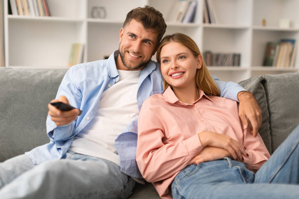 Happy Young Couple Watches Television Show, Switching Channels With Remote Control, Enjoying Movies While Hugging And Relaxing On Sofa At Home. Domestic Leisure And Fun On Weekend - Photo, Image