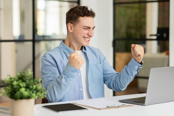 Happy excited millennial caucasian man manager rise fists, celebrate win, success business, look at computer at table in modern coworking office. Good work, professional career - Photo, Image
