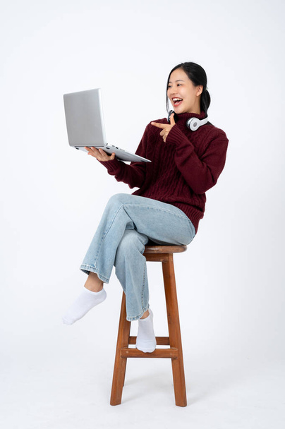 Νεαρή χαρούμενη Ασιάτισσα με ζεστά ρούχα δείχνει το δάχτυλό της σε μια οθόνη laptop, απολαμβάνοντας στα μέσα κοινωνικής δικτύωσης, κάθεται σε μια καρέκλα σε απομονωμένο λευκό φόντο. - Φωτογραφία, εικόνα