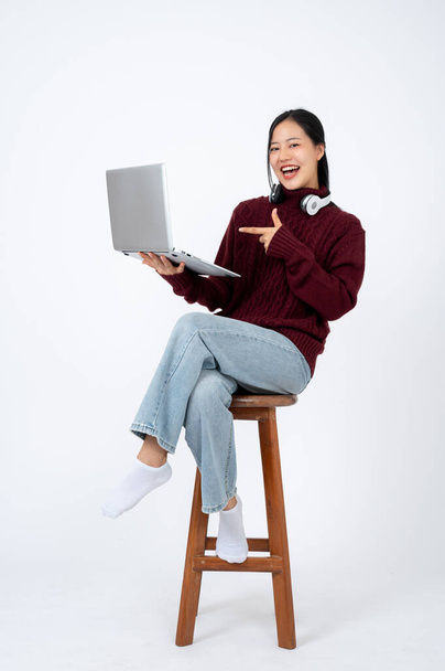 Joven mujer asiática alegre en ropa acogedora está apuntando con el dedo a la pantalla de un ordenador portátil, disfrutando en las redes sociales, sentado en una silla sobre un fondo blanco aislado. - Foto, Imagen