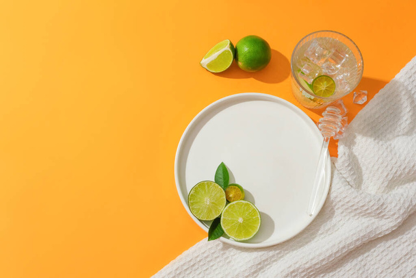 Blick von oben auf Zitronen-, Kumquat- und grüne Blätter auf einem runden Keramikteller, einem mit Eis und einem weißen Tuch gefüllten Glas Wasser. Freiraum für Werbung. - Foto, Bild