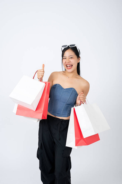 Μια χαρούμενη και κομψή Ασιάτισσα με μοντέρνα ρούχα κρατάει τις τσάντες της, στέκεται πάνω σε ένα απομονωμένο λευκό φόντο. εφαρμογή για ψώνια, online αγορές, μόδα και τρόπος ζωής - Φωτογραφία, εικόνα