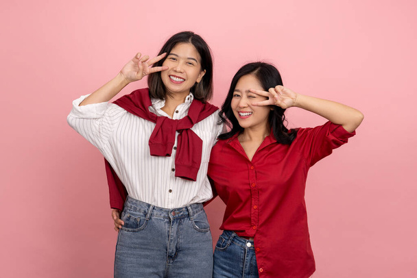 Twee gelukkige Aziatische vrouwelijke vrienden tonen v teken op roze achtergrond. en kijkend naar de camera - Foto, afbeelding