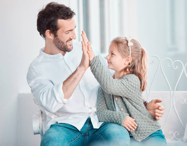 Father, girl child and happy with high five, smile and hug with care, love and bonding on chair in home. Dad, kid and cheers with success, support and motivation at family house for celebration. - Photo, Image