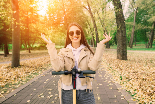 Young beautiful stylish woman riding electric scooter in autumn park, ecological transport - Photo, Image