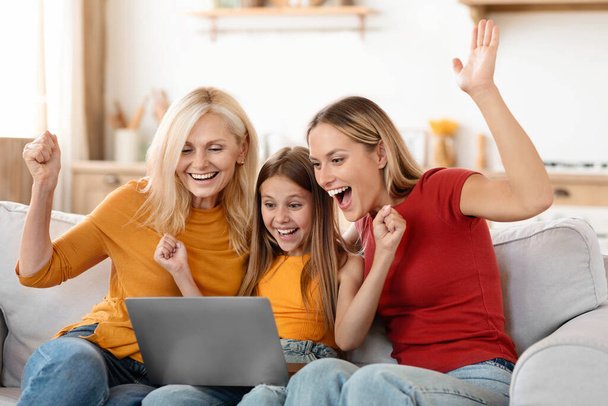 Grand-mère émotionnelle, mère et fille utilisant un ordinateur portable à la maison, assis sur le canapé dans le salon, regardant l'écran d'ordinateur, levant les mains et riant, jouant en ligne, amusez-vous - Photo, image