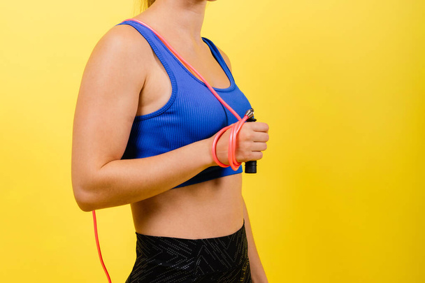 Portrait of gentle muscular woman holding skipping rope on her neck over yellow background - Photo, Image