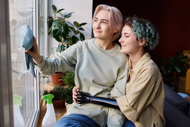 Urocza lesbijska para wykonuje prace domowe w nowym mieszkaniu podczas przeprowadzki, wycierają okno z kurzu - Zdjęcie, obraz