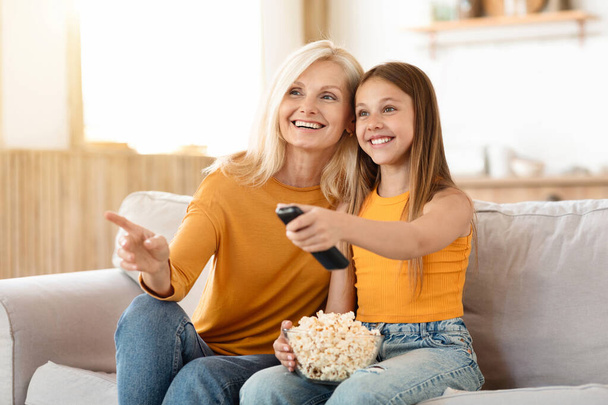 Glückliche Enkelin und Großmutter beim Kinobesuch zu Hause. Fröhliches Teenie-Mädchen genießt die Zeit mit ihrer Oma, wählt Cartoon im Fernsehen, isst Popcorn, kopiert Raum - Foto, Bild