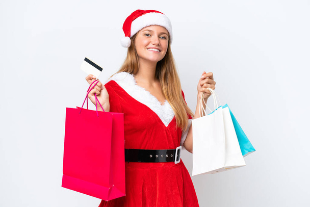 Joven mujer caucásica vestida como mama noel aislada sobre fondo blanco sosteniendo bolsas de compras y una tarjeta de crédito - Foto, imagen