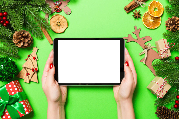 Top view of woman holding tablet in her hands on green background made of Christmas decorations. New Year holiday concept. Mockup. - Photo, Image