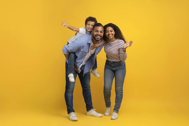 Vrolijke Afro-Amerikaanse familie van drie plezier hebben samen over gele achtergrond, gelukkige jonge zwarte ouders en hun pretiaan zoon lachen en glimlachen op camera, volledige lengte schot, kopieerruimte - Foto, afbeelding
