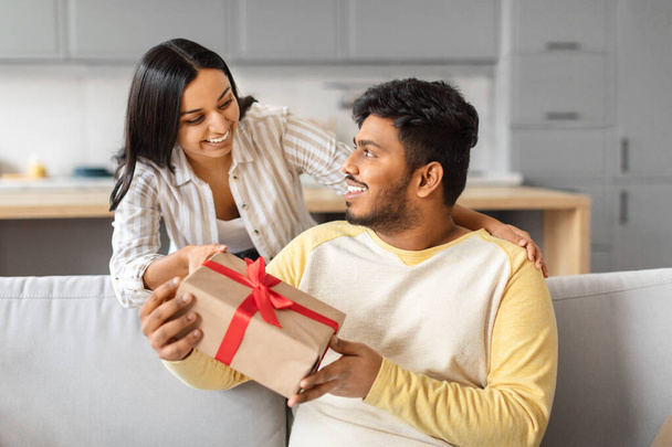 Amante esposa india sorprendiendo a su marido con regalo en casa, saludándolo con cumpleaños o aniversario, feliz mujer oriental dando caja de regalo a hombre emocionado, celebrando el día de San Valentín juntos - Foto, imagen