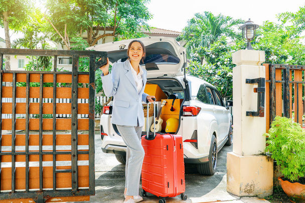 Viszlát öreg ház: boldog ázsiai üzletasszony csomagolja az előkészületeket csomagok autóba készül mozog, nyomja piros bőrönd áll a bejárati ajtó néz kamera mosolygós utazás vezetés áthelyezés. - Fotó, kép