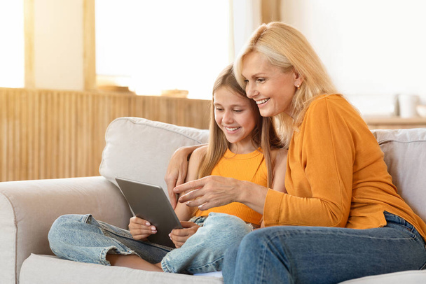 Милая девочка-подросток весело проводит время дома со своей бабушкой, сидя на диване в гостиной, обнимая и используя цифровой планшет, покупки в Интернете, используя хорошее мобильное приложение, копировать пространство - Фото, изображение
