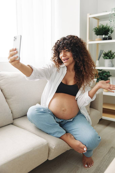 Terhes nő blogger ül a kanapén otthon, és képeket készít magáról a telefonon, selfie és videó hívás, konzultáció az orvossal az interneten, a terhesség kezelése. Kiváló minőségű fénykép - Fotó, kép