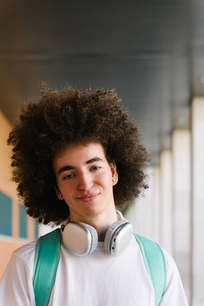 Vertikální portrét chladného mladého studenta s kudrnatými vlasy, usmívajícího se do kamery stojící na kampusu - Fotografie, Obrázek