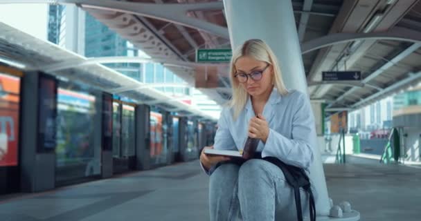 Žena, která čte knihu na stanici metra. Žena zavírá knihu, rozhlíží se, sedí u vchodu do metra. Mladá studentka studující literaturu čtení knihy mimo venkovní - Záběry, video