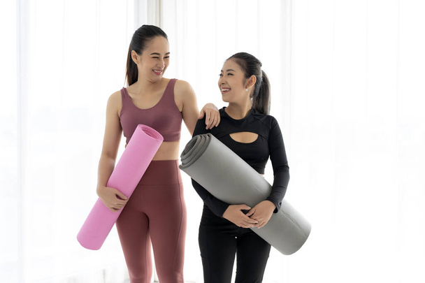Two fit Asian women stand side by side, holding rolled yoga mats and smiling at the camera. They are ready to start their class and look happy and excited. - Foto, Imagem