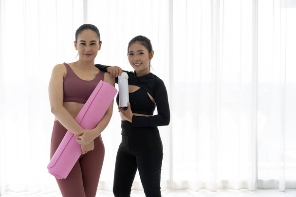 Two fit Asian women stand side by side, holding rolled yoga mats and smiling at the camera. They are ready to start their class and look happy and excited. - 写真・画像