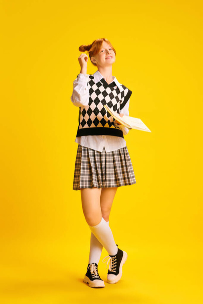 Faull-length portrait of adorable red haired girl, dressed retro fashion outfit reading book against yellow background. Concept of youth, education, fashion, study, knowledge, - Photo, Image