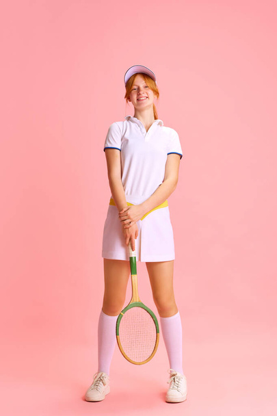 Pełnowymiarowy portret pięknej, uśmiechniętej młodej rudowłosej kobiety, profesjonalnej tenisistki pozującej z rakietą na pastelowym różowym tle. Pojęcie sportu zawodowego, rekreacji, hobby. - Zdjęcie, obraz