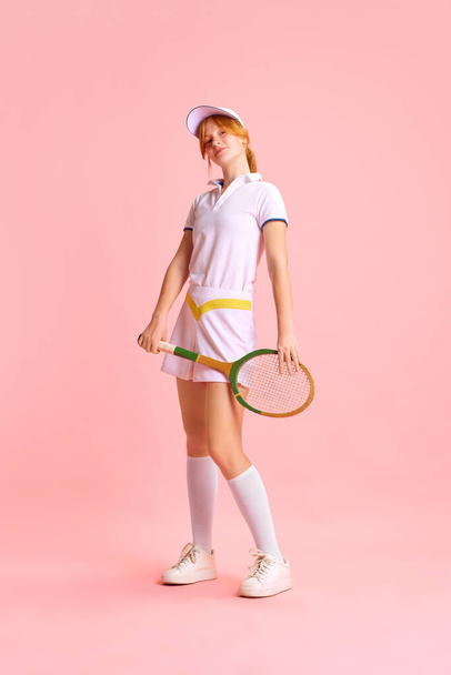 Piękna, uśmiechnięta młoda rudowłosa kobieta, profesjonalny tenisista w białym mundurze pozujący z rakietą na pastelowym różowym tle. Pojęcie sportu zawodowego, rekreacji, hobby. - Zdjęcie, obraz