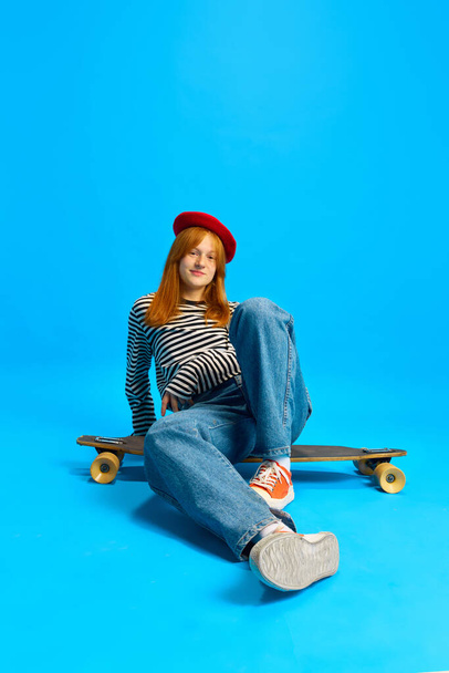 Beautiful young lady, student dressed retro fashion outfit in red beret posing sitting skateboard against blue studio background. Concept of human emotions, fashion, style, sales season, youth culture - Photo, Image