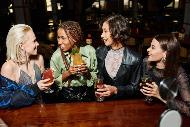 χαμογελαστές και κομψές πολυφυλετικές φιλενάδες με ποτήρια κοκτέιλ που μιλάνε στο μπαρ, ζωντανή ατμόσφαιρα - Φωτογραφία, εικόνα