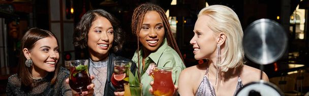 sonrientes y elegantes amigas multiculturales sosteniendo copas de cócteles en el bar, pancarta horizontal - Foto, imagen