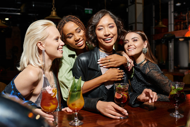 happy african american woman embracing asian girlfriend in cocktail bar, hen party at nighttime - Photo, Image
