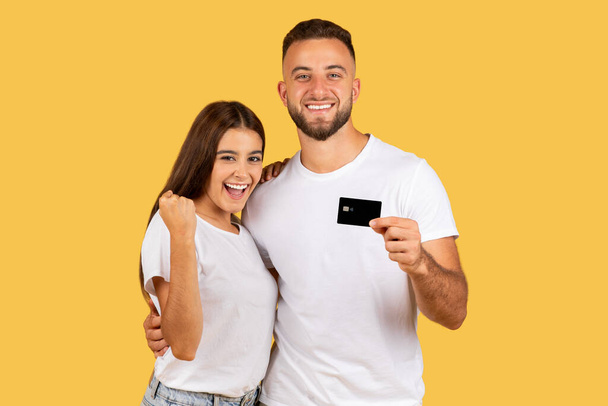 Glad young european family in white t-shirts, with credit card hugs, celebrate success, win, isolated on yellow studio background. Ad and offer, win, sale recommendation, shopping, cashback - Photo, Image
