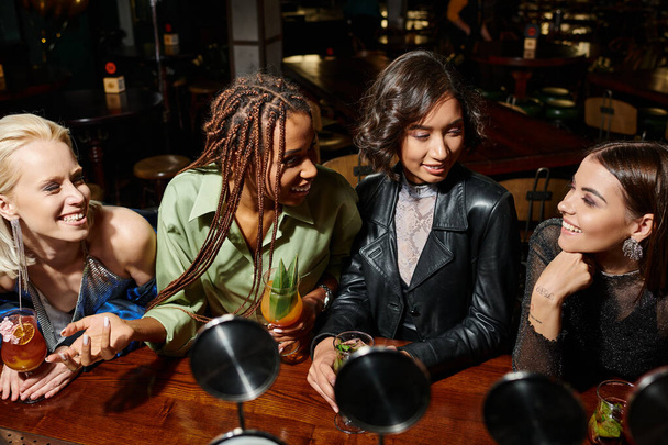 iloinen monikansallinen naispuoliset ystävät puhuvat ja hymyilevät lähellä cocktail lasit, kana puolue baarissa - Valokuva, kuva